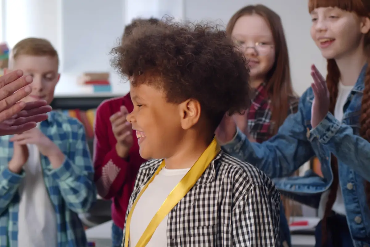 Smiling child wearing a medal while classmates applaud around him.