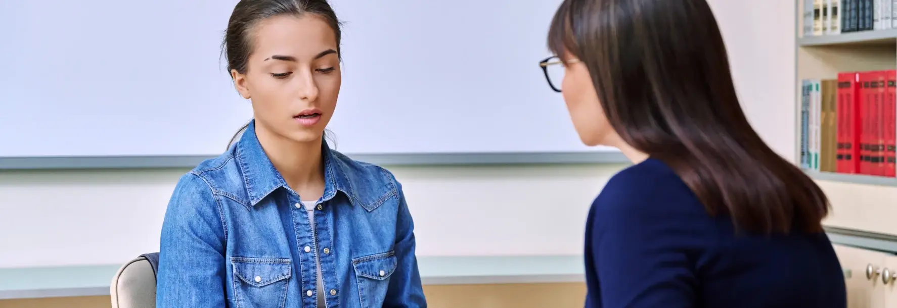 Young woman in a denim shirt speaking with a counselor during a private session in a quiet, book-filled office setting.