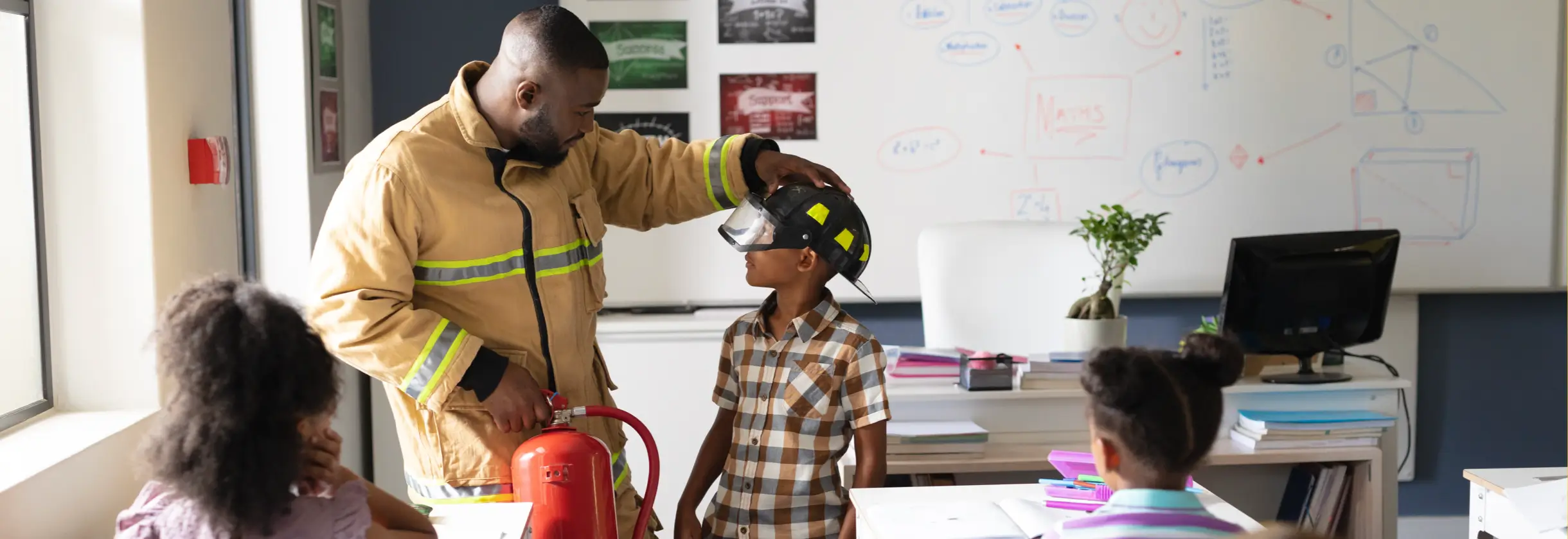 Fireman visiting students in a classroom and putting his helmet on a student.