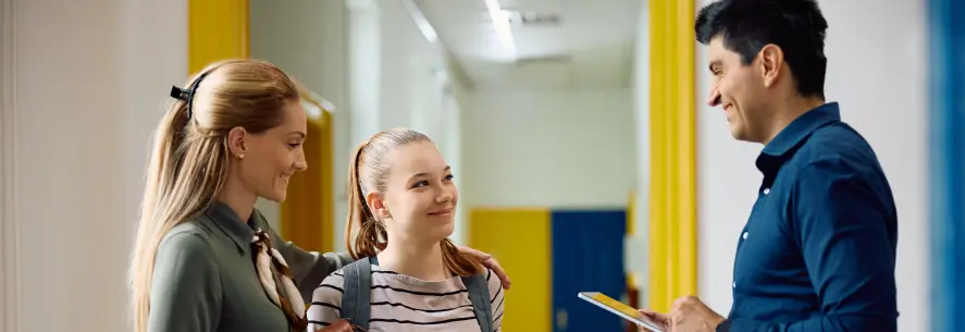 Male teacher talking to a female student with her mother in a school hallway.