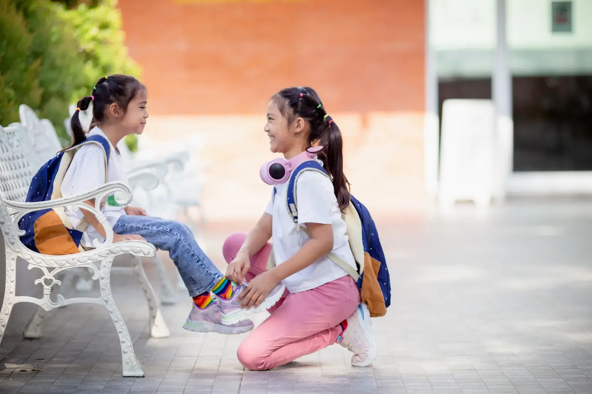 Two school-age girls with backpacks smiling and chatting as one ties the other’s shoe on a bench outdoors.